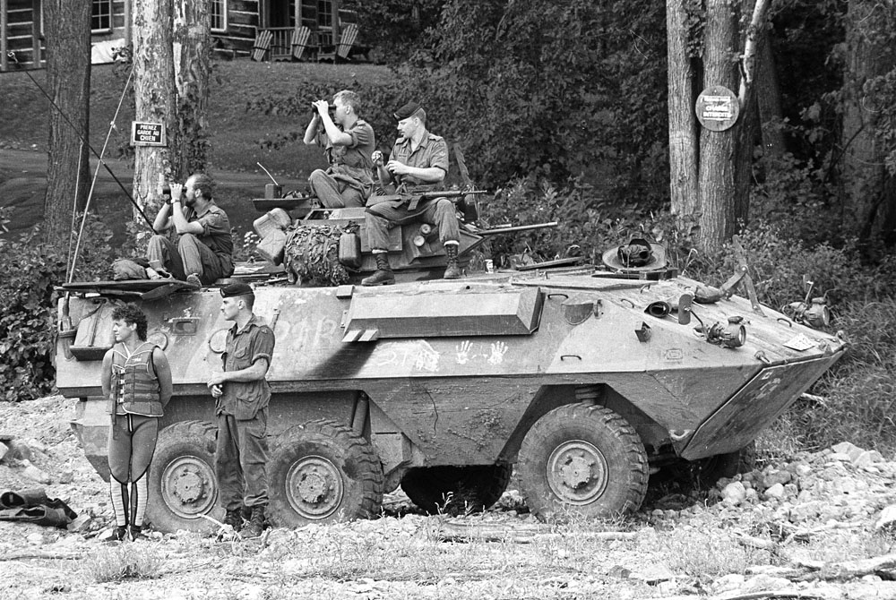 A Grizzly APC with a few soldiers around in front of some trees and a house in the background