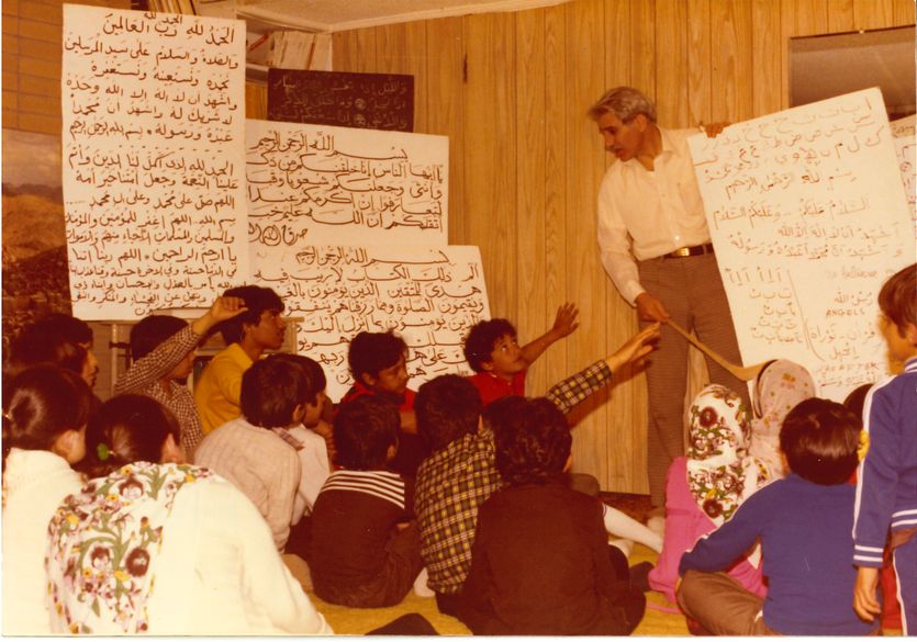A man in a white shirt alongside some white signs with letters teaches a room full of children